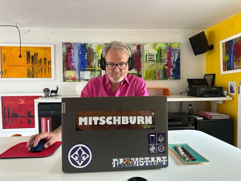 Mitsch Schulz am Laptop mit MITSCHBURN-Aufkleber, im Hintergrund seine Bilder