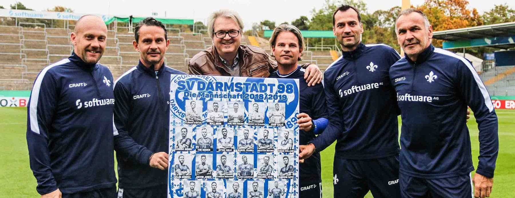 Trainerteam des SV Darmstadt 98 mit Mitsch Schulz und dem Mannschaftsposter im Comicstil auf dem Rasen